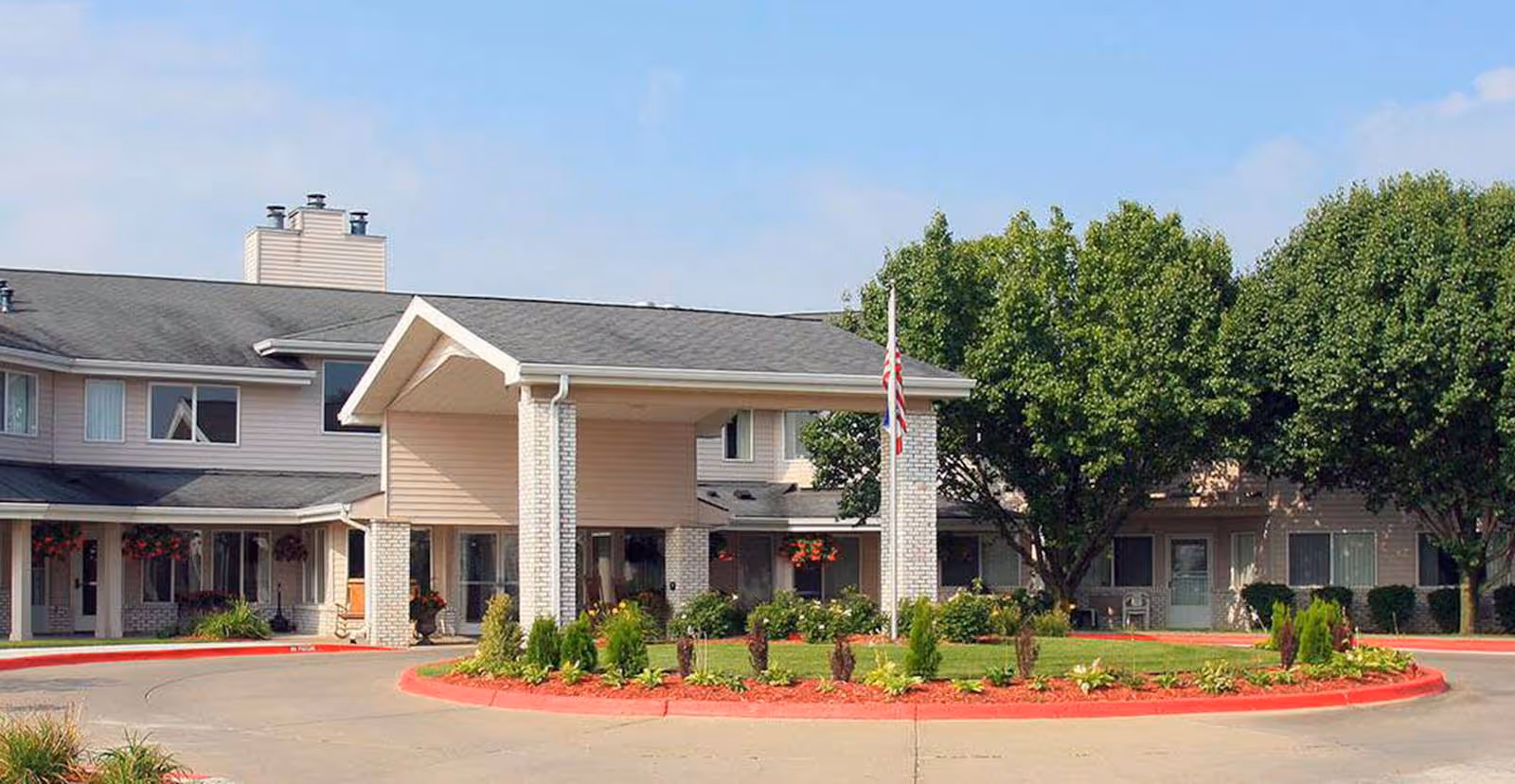 Front entrance of a senior living building with a porte-cochère, circular driveway, landscaped island and an American flag.