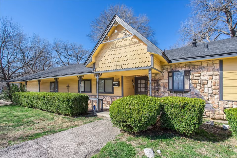 Front exterior of a yellow single-story house with stone accents, a covered porch, trimmed hedges and a walkway under a clear blue sky.