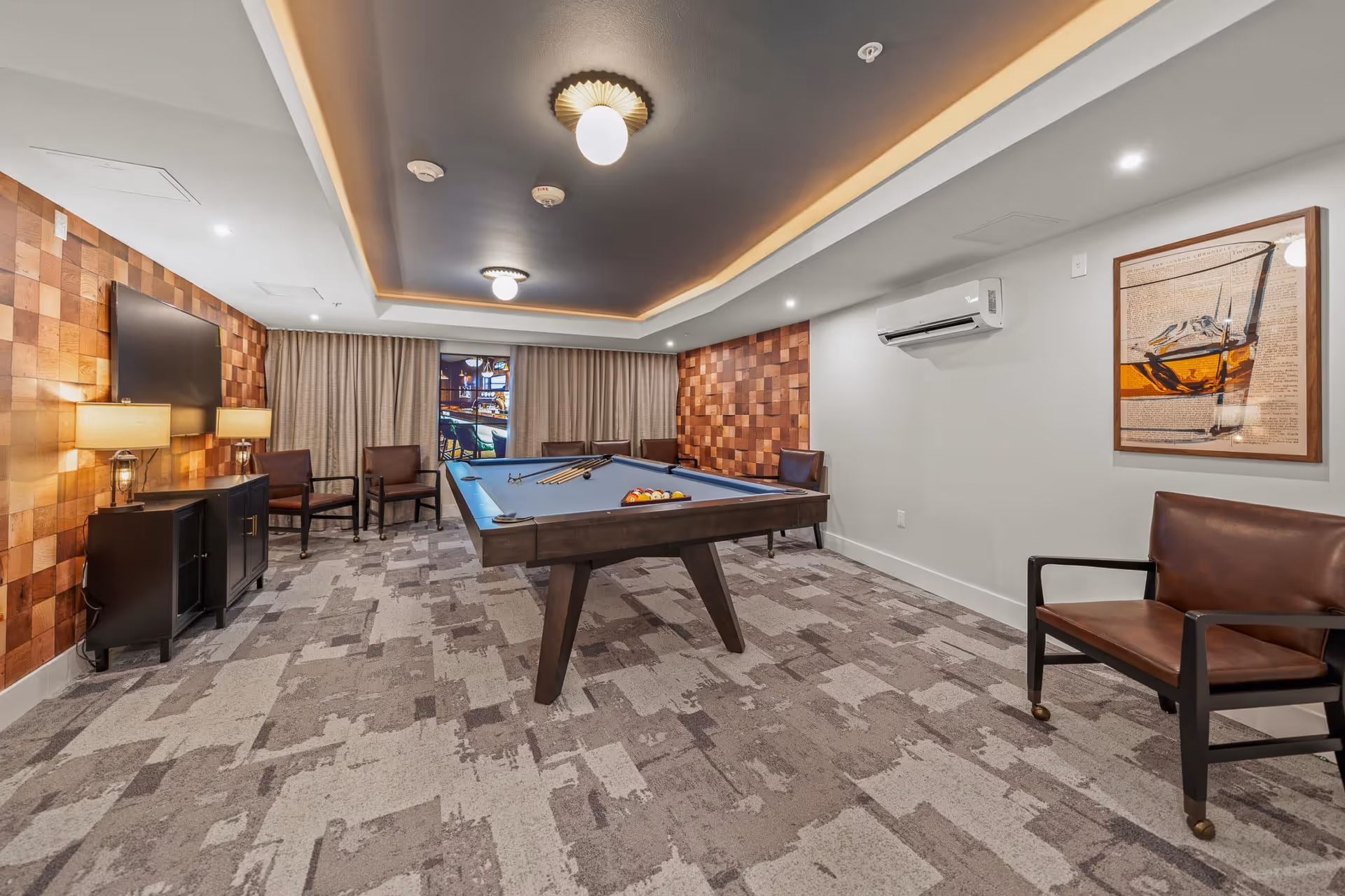 A modern game room with a pool table in the center, surrounded by several brown leather chairs. The room features a patterned carpet, wood-paneled accent walls, a mounted flat-screen TV, two table lamps on a black cabinet, and a framed artwork on the wall. The ceiling has recessed lighting and decorative light fixtures, and there is an air conditioning unit mounted on the wall.