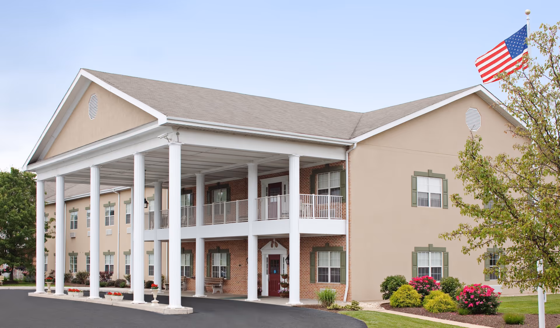 Exterior view of a two-story senior living facility building with beige walls, green window shutters, and a covered entrance supported by white columns. There is an American flag on a flagpole to the right, along with landscaped bushes and flowers.