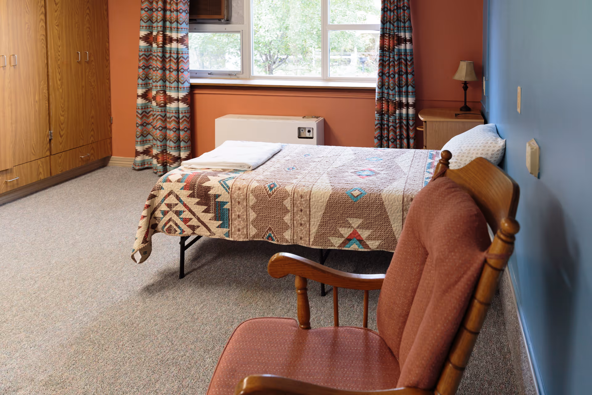 A cozy bedroom with a single bed covered in a patterned quilt, a wooden rocking chair with a cushioned seat, a wooden wardrobe, a small bedside table with a lamp, and a window with patterned curtains letting in natural light.