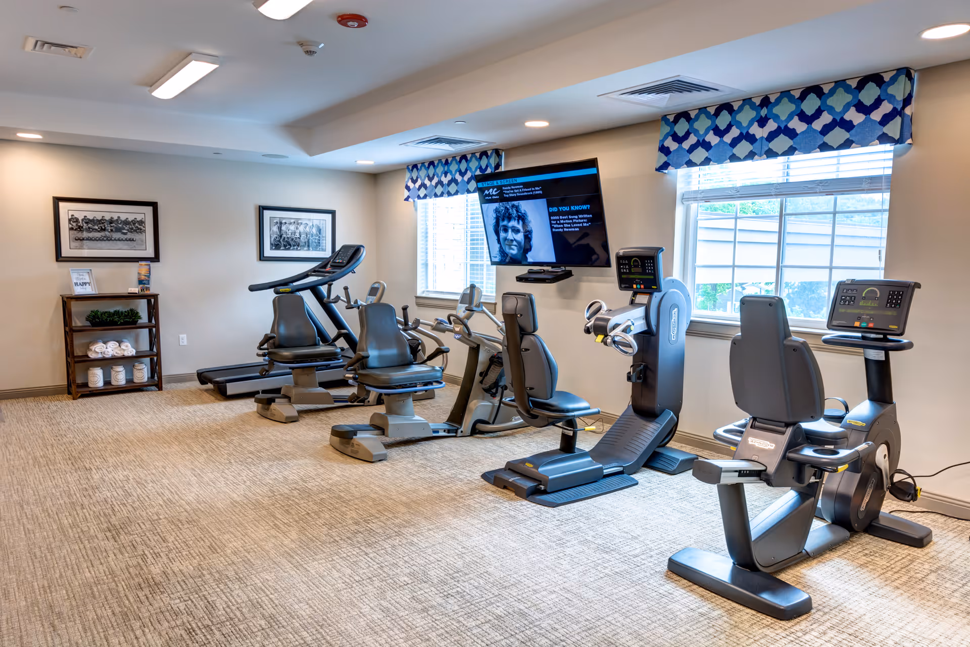 A senior living facility exercise room with several pieces of cardio equipment including recumbent bikes and a treadmill. The room has beige carpet, two windows with blue patterned valances, framed black and white photos on the wall, and a TV mounted between the windows.