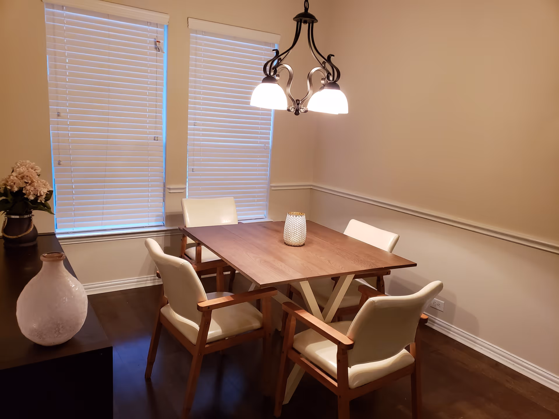 A dining room with a wooden table surrounded by four cream-colored cushioned chairs with wooden arms. Two windows with closed white blinds are on the wall behind the table. A dark wooden sideboard with a white vase and a flower arrangement is on the left side. A chandelier with three lights hangs above the table.