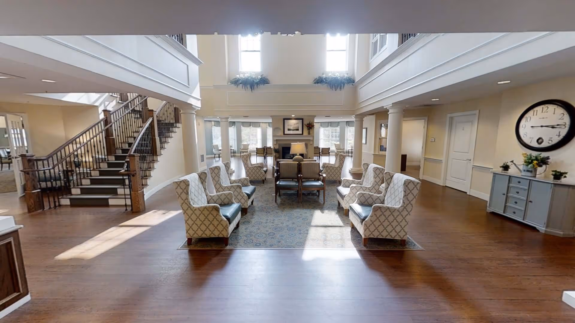 Spacious two-story communal living area with armchairs arranged around a rug, a staircase to the left, and a large wall clock on the right.