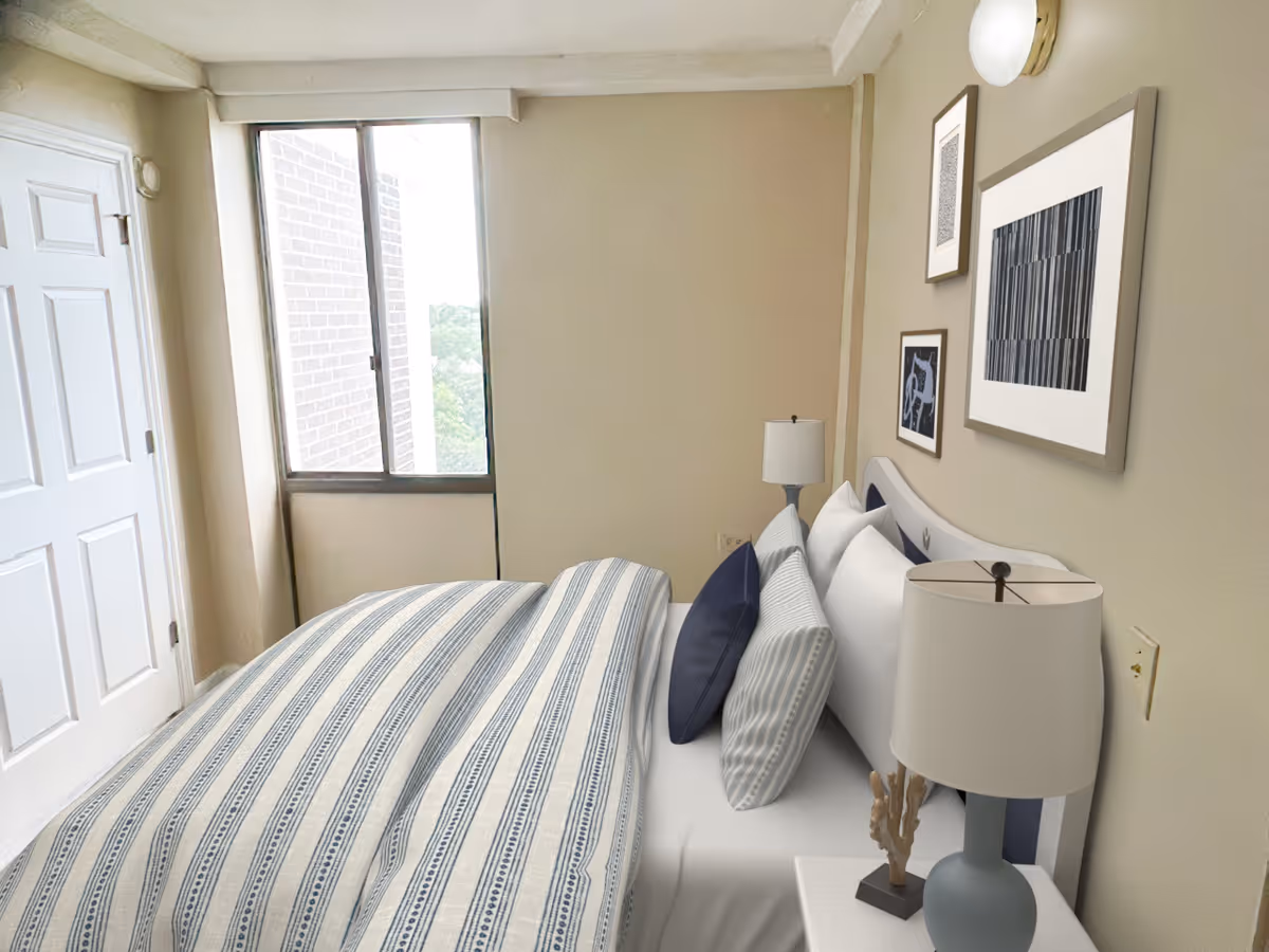 A bedroom with a neatly made bed featuring striped bedding and multiple pillows. There are two bedside tables with lamps, and framed artwork is hung on the wall above the bed. A window with a view of a brick wall and greenery is visible, along with a closed white door.