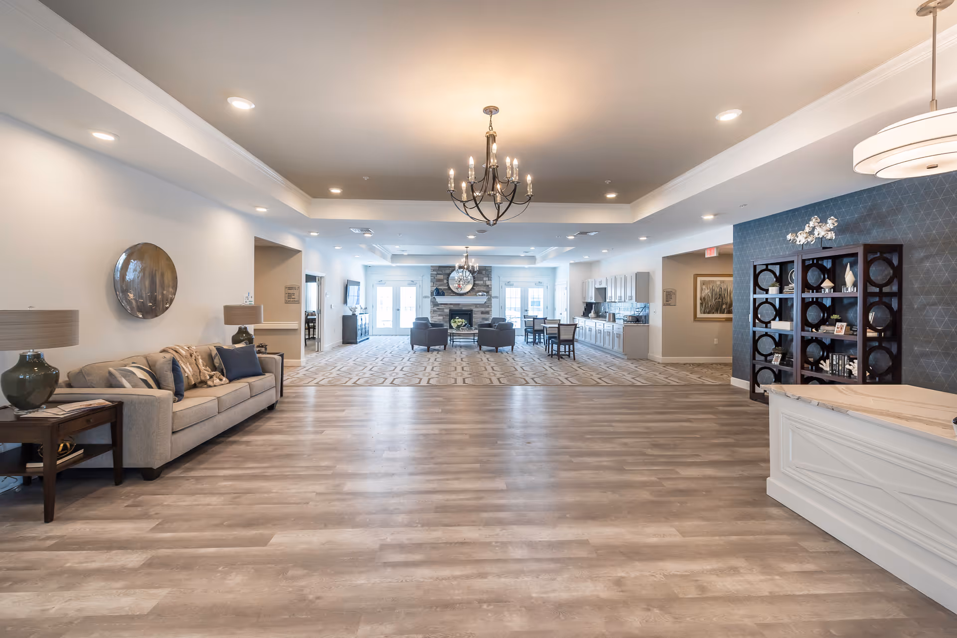Spacious and well-lit senior living facility common area with a chandelier, a beige sofa with cushions, side tables with lamps, a decorative wall mirror, a seating area with armchairs around a stone fireplace, a dining table with chairs, and a reception desk with shelving on the right side.