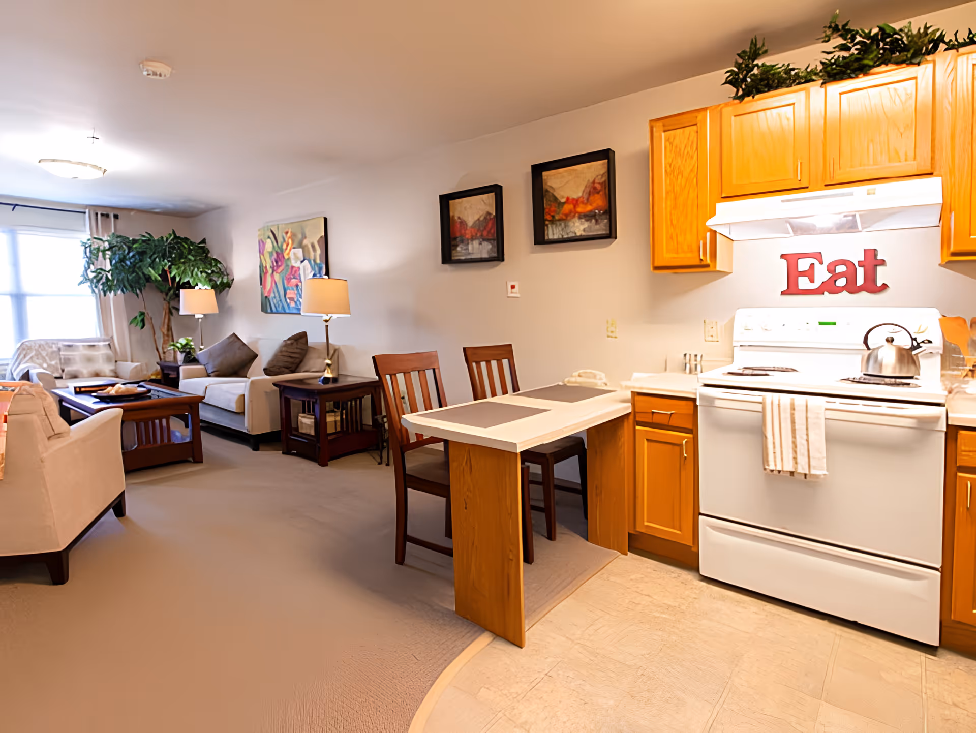 Open-plan living area and kitchenette with sofas, a small dining table, potted plant, and a stove with a red "Eat" sign above it.