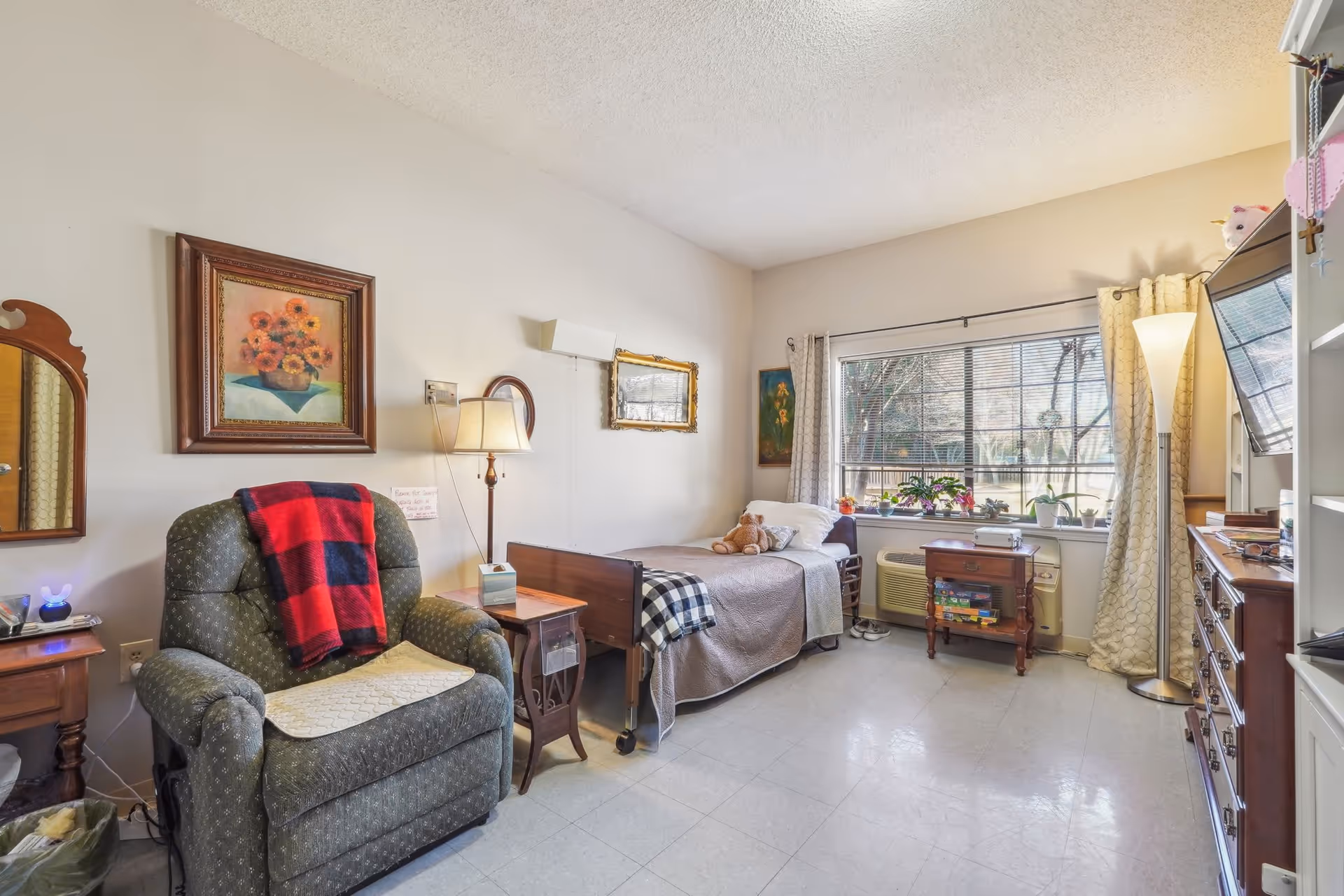 A cozy senior living bedroom with a single bed covered in a beige quilt and a teddy bear on top. Next to the bed is a wooden side table with a lamp and a green upholstered recliner chair with a red and black checkered blanket draped over it. The room has a large window with blinds and several potted plants on the windowsill. There is a tall floor lamp near the window, a wooden dresser with a TV mounted above it, and framed artwork on the walls. The floor is tiled and the room is well-lit with natural light.