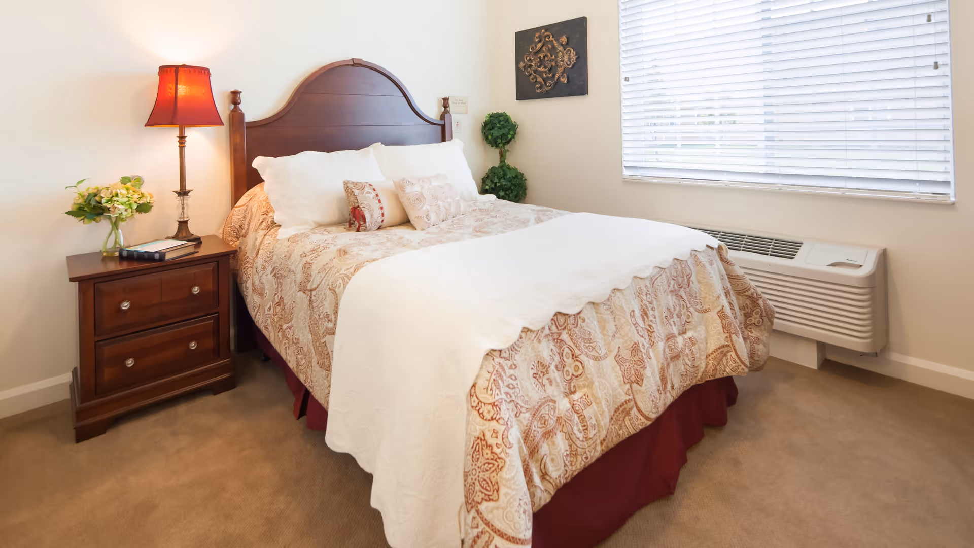 A neatly made bed with a patterned bedspread and white pillows in a bedroom. Next to the bed is a wooden nightstand with two drawers, a table lamp with a red shade, a book, and a small vase of flowers. A window with white blinds and an air conditioning unit are visible on the right wall. A decorative wall piece and a small green plant are also present in the room.