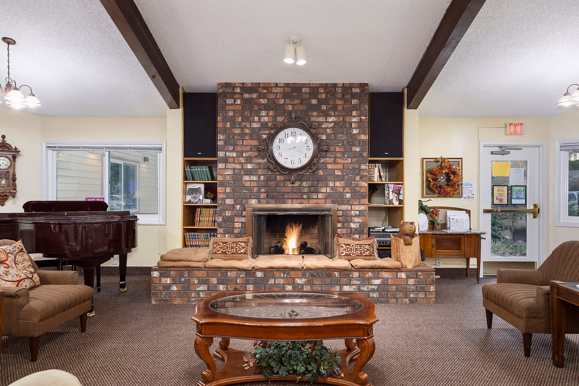 Cozy common room in a retirement community featuring a central brick fireplace with a clock above, cushioned seating, a grand piano, and a glass-topped coffee table.