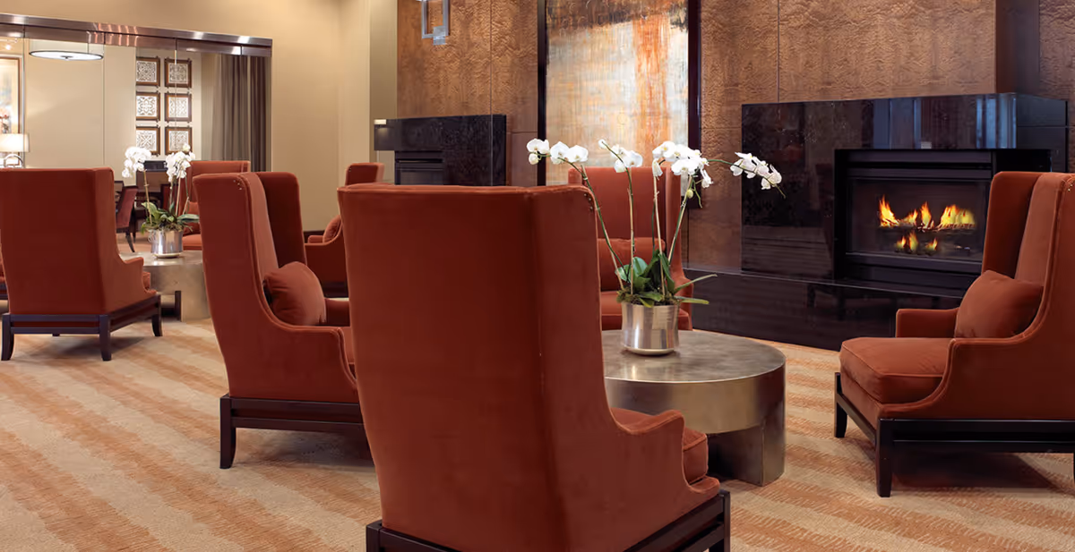 A cozy lounge with rust-colored armchairs arranged around a round coffee table and a lit fireplace.