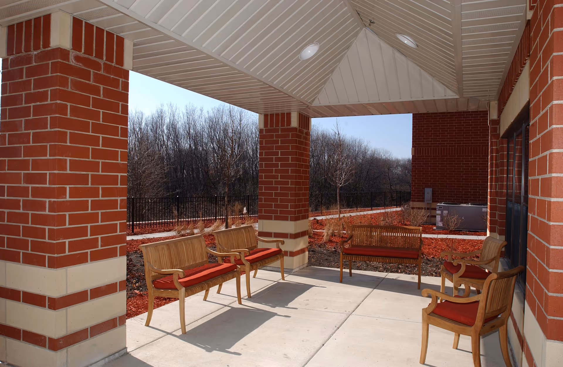 Covered outdoor seating area with wooden benches and chairs with red cushions, surrounded by brick pillars and a landscaped garden with bare trees in the background.