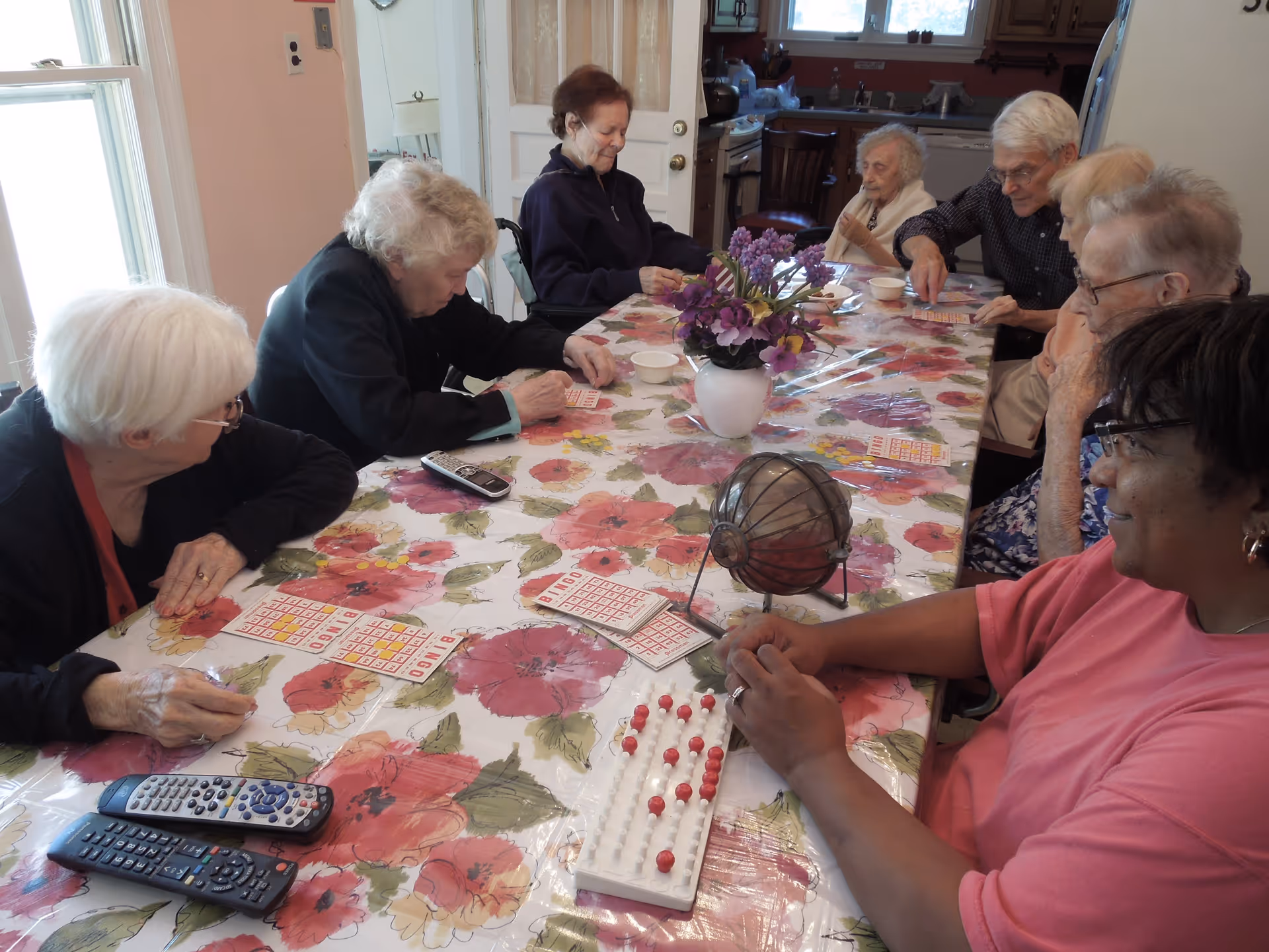 A group of elderly people sitting around a floral-patterned table playing bingo. There is a bingo cage and bingo cards on the table, along with a vase of purple flowers. The setting appears to be a cozy indoor room with windows and kitchen furniture in the background.