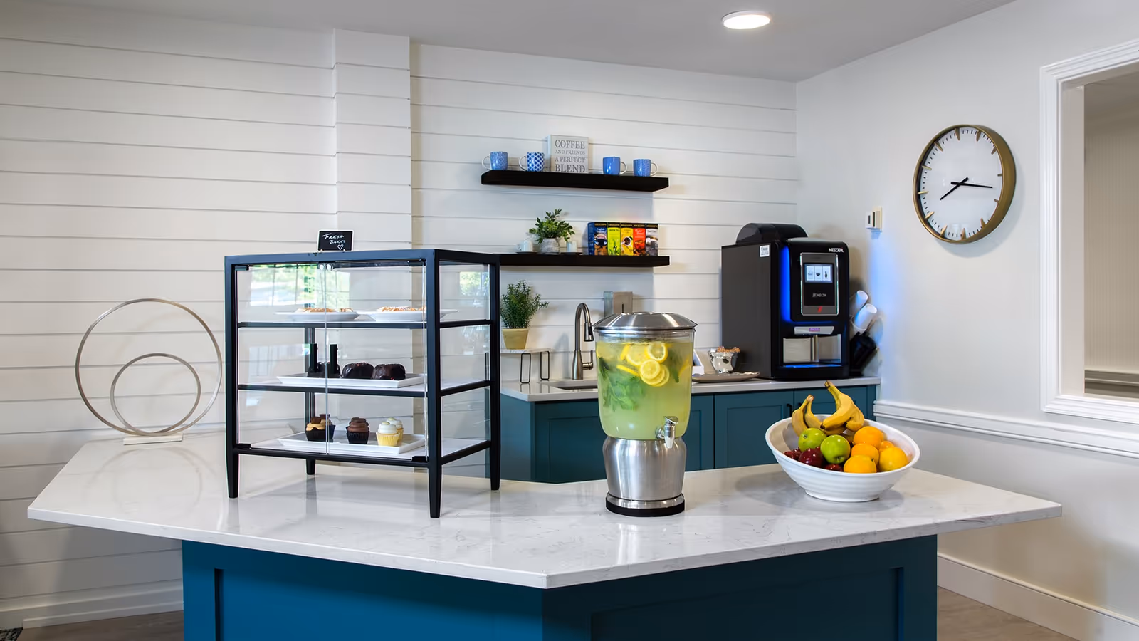 A modern refreshment area with a white marble countertop and teal cabinetry. On the counter, there is a glass display case with assorted cupcakes and pastries, a large beverage dispenser filled with lemon and mint infused water, and a white bowl containing bananas, apples, oranges, and grapes. Behind the counter, there is a coffee machine, a sink, two floating shelves with coffee mugs, plants, and coffee packets. A round wall clock is mounted on the white wall to the right.