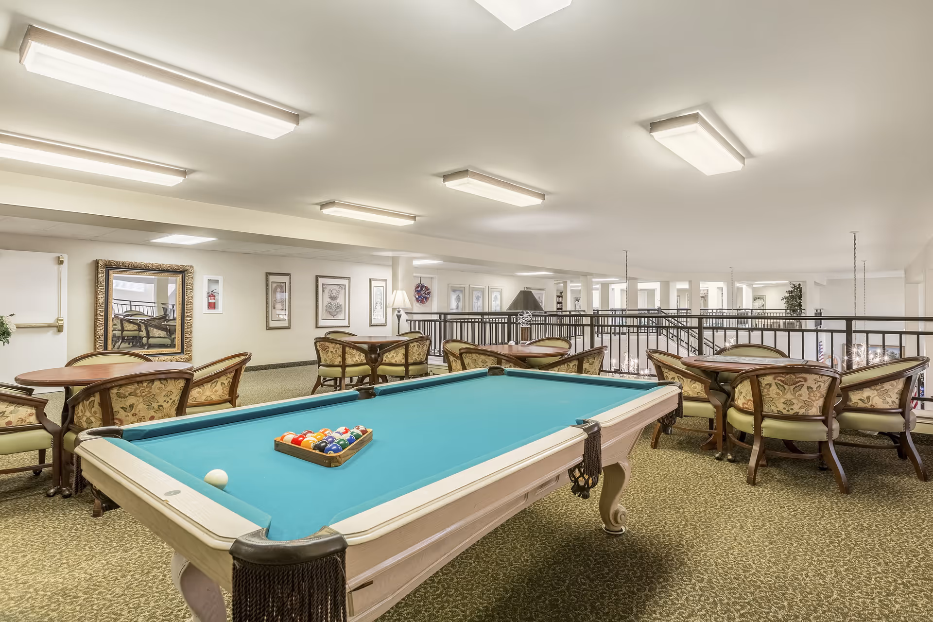 A senior living community game room with a pool table in the foreground and several round tables with upholstered chairs arranged around the room. The room has carpeted floors, framed artwork on the walls, and bright overhead lighting. A railing overlooks a lower level with chandeliers visible below.