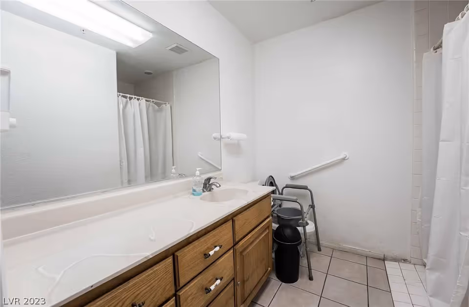 Bathroom with a large mirror above a wooden vanity with a sink and soap dispenser. There is a white shower curtain on the right side, a grab bar on the wall, and a walker with a seat and a black trash can next to it. The floor is tiled.