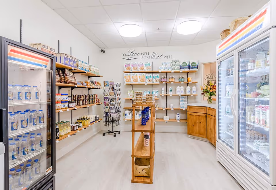 Interior view of a small grocery or convenience store area within a facility, featuring shelves stocked with various food items and beverages. There are refrigerated display cases on both sides filled with drinks, dairy products, and other perishable goods. Wooden shelves on the back wall hold packaged foods, and a small wooden island shelf is in the center. The wall has a decorative quote that reads 'To Live Well is to Eat Well.' The space is brightly lit with ceiling lights and has a clean, organized appearance.