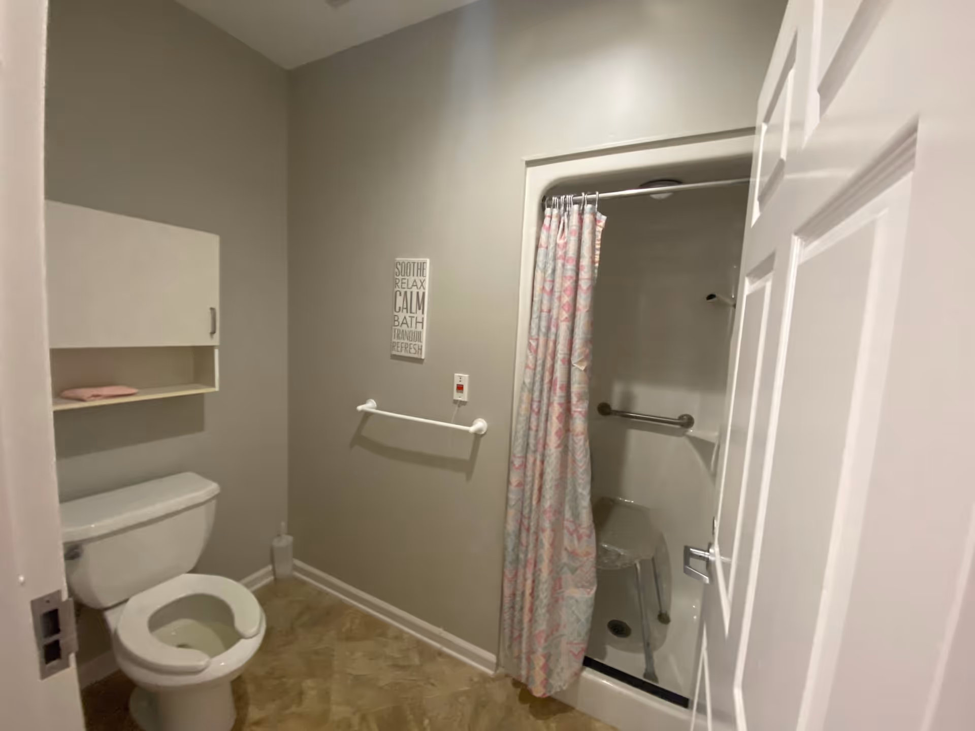 Small accessible bathroom with a toilet, wall cabinet, towel bar, and shower with a curtain, grab bar, and stool.