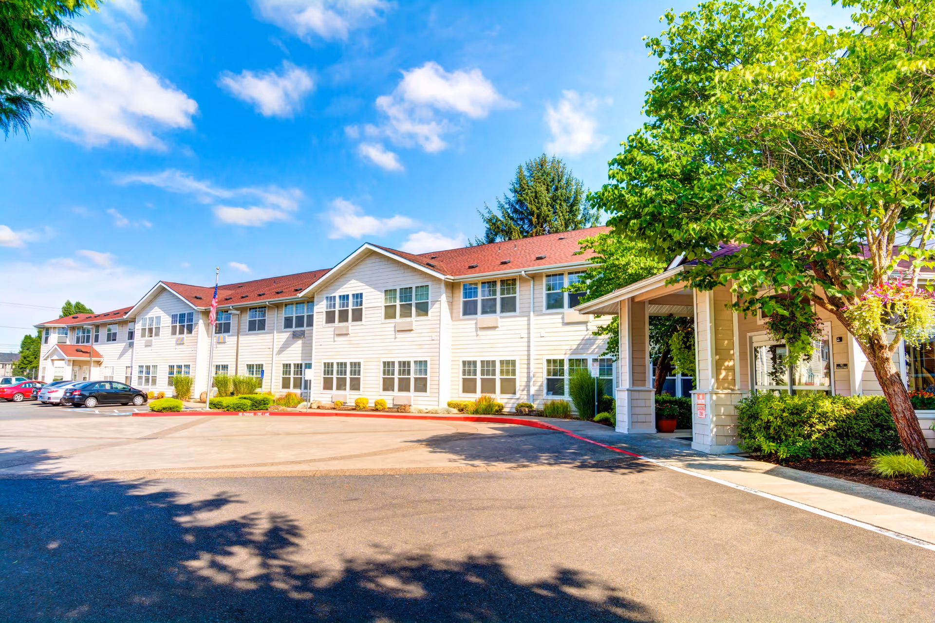 Exterior view of a two-story senior living facility building with beige siding and a red roof under a blue sky with scattered clouds. There are several parked cars in front of the building, a flagpole with an American flag, and a covered entrance with trees and shrubs around it.