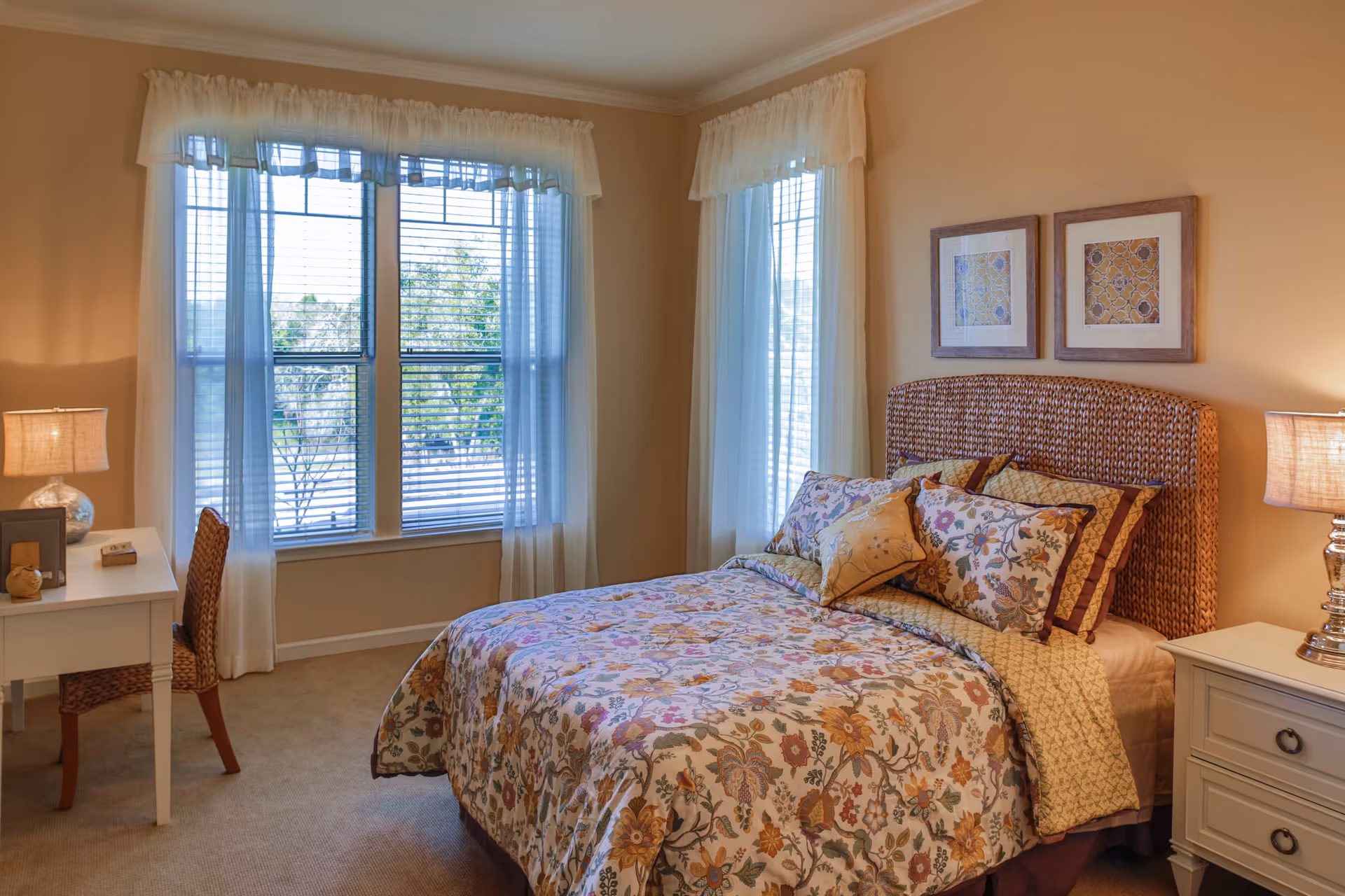 A cozy bedroom with a bed featuring a floral patterned bedspread and multiple pillows. The room has two large windows with sheer white curtains allowing natural light to enter. There is a white desk with a wicker chair and a lamp on the left side, and a white nightstand with a lamp on the right side. Two framed artworks hang on the wall above the bed.