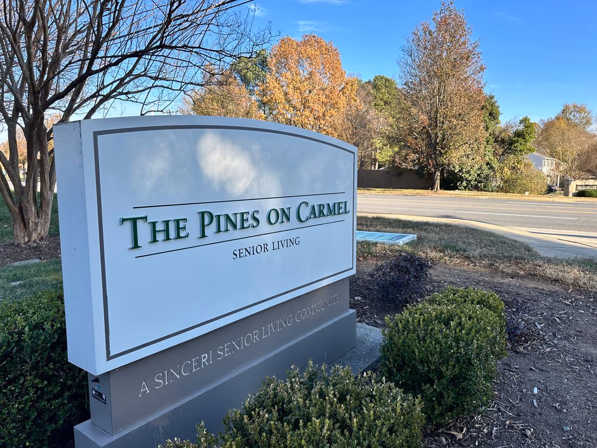 Entrance sign for The Pines on Carmel senior living community beside a road with trees and shrubs.