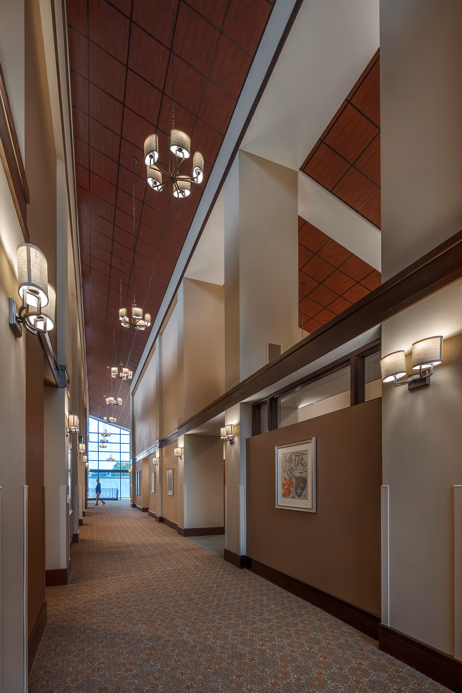 A long, carpeted hallway in a senior living facility with high ceilings and multiple modern light fixtures hanging from a wooden-paneled ceiling. The walls are painted in neutral tones with framed artwork and wall sconces evenly spaced along the corridor. At the far end, large windows allow natural light to enter, and a person is visible walking near the windows.
