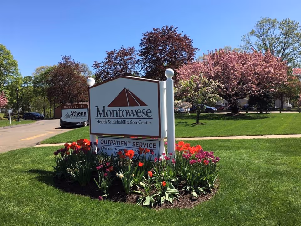Outdoor view of the Montowese Health & Rehabilitation Center sign surrounded by a flower bed with red and purple tulips, green grass, and trees with pink blossoms under a clear blue sky.
