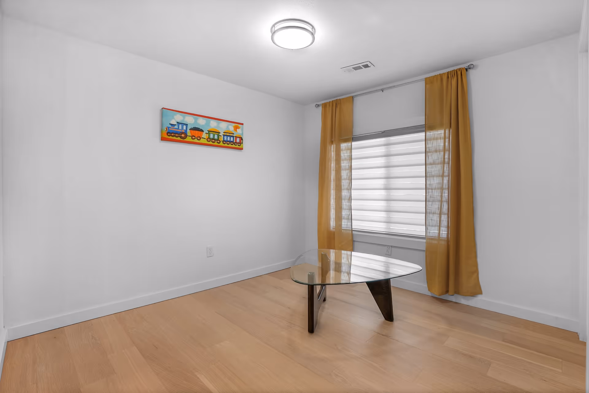 A minimalist room with light wooden flooring, white walls, a glass coffee table with dark wooden legs, a window with white blinds and mustard yellow curtains, and a colorful painting of a train on the wall.
