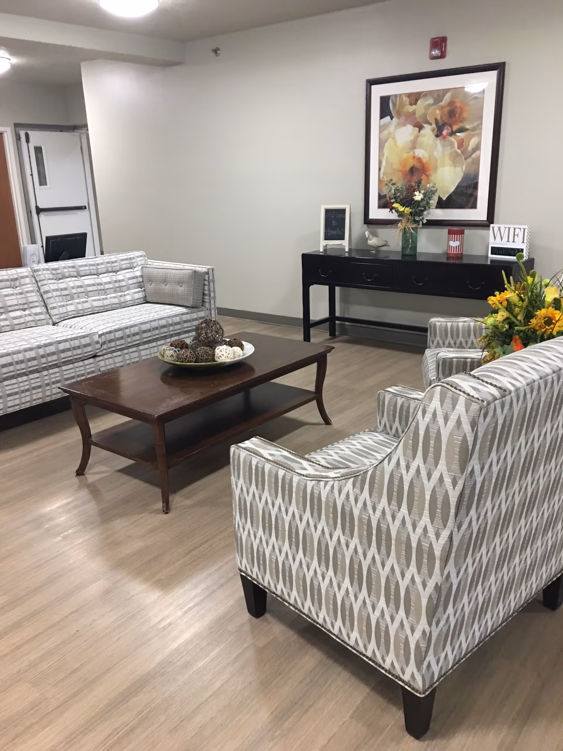 A cozy living room area with patterned sofas and armchairs arranged around a wooden coffee table with decorative balls. A black console table against the wall holds a vase with flowers, a small chalkboard, a bird figurine, and a sign displaying the WiFi network name. Above the console table hangs a large framed floral painting. The room has light-colored wood flooring and neutral-colored walls.