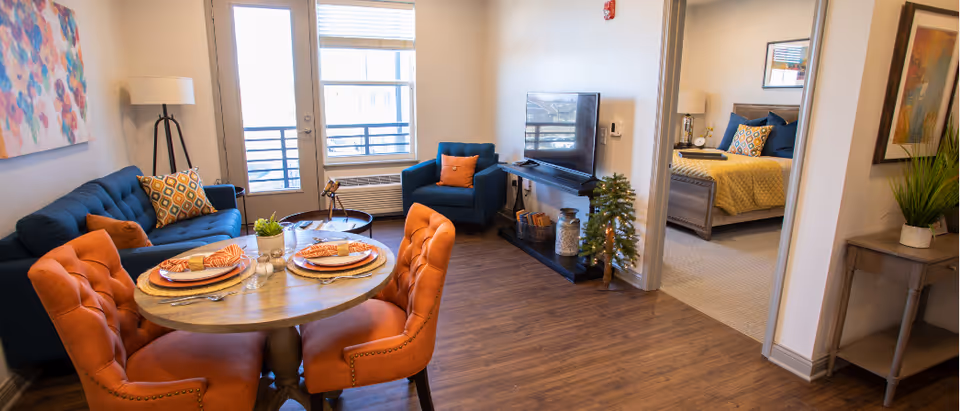 Open living and dining area of a furnished apartment with a round table and orange chairs, blue sofa and armchair, TV stand, and a bedroom visible through a doorway.