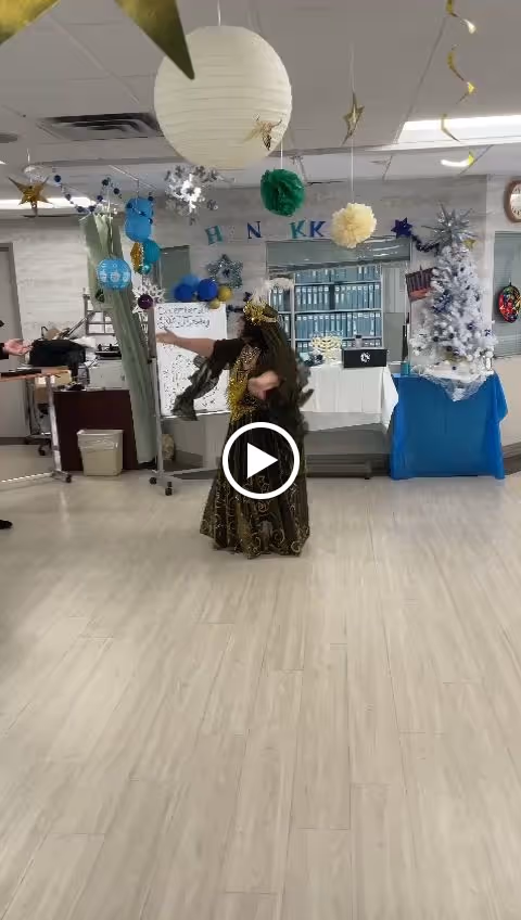 A person dressed in a decorative costume is dancing in a decorated room with hanging paper lanterns, stars, and other festive ornaments. There is a white Christmas tree with blue and silver decorations on the right side, and a table covered with a white cloth and blue skirt in the background. The room has light-colored wooden flooring and a white ceiling with fluorescent lights.