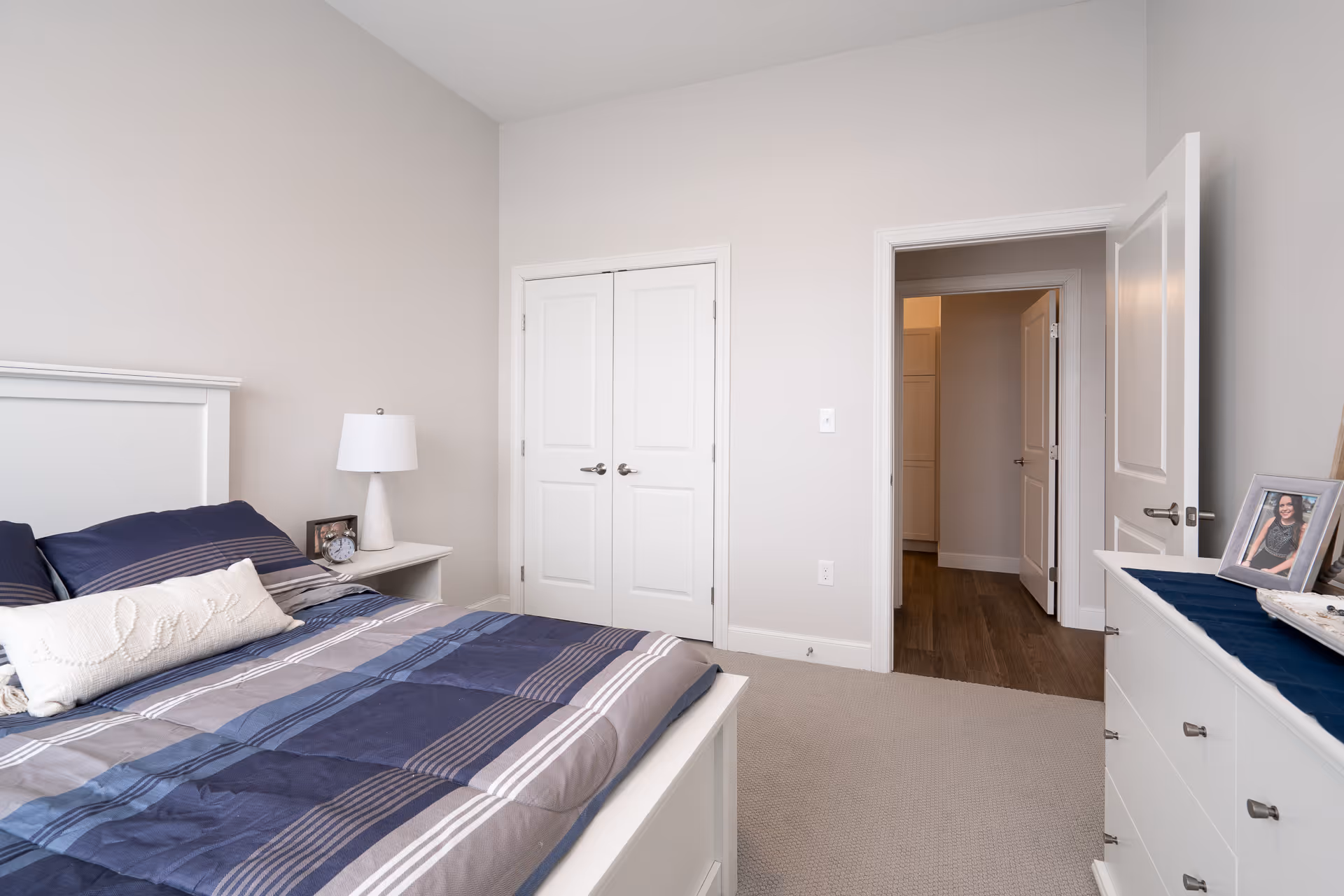 A neatly arranged bedroom with a white bed frame and a blue and gray striped comforter. There is a white nightstand with a white lamp and a framed photo next to the bed. Across from the bed is a white dresser with framed photos on top. The room has beige walls, a beige carpet, and two white doors, one leading to a hallway and the other a closet.