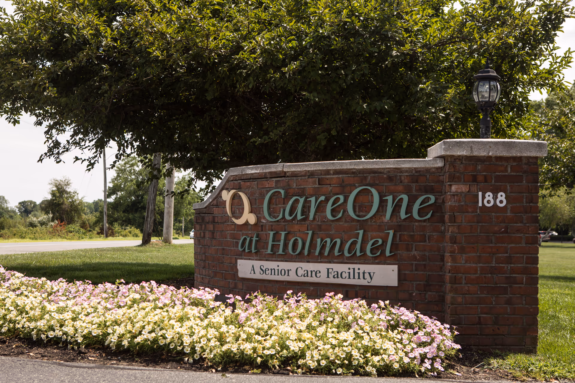A brick sign for CareOne at Holmdel, a senior care facility, surrounded by green grass, trees, and a flower bed with white and light purple flowers. The sign includes the address number 188 and a small lamp post on top.