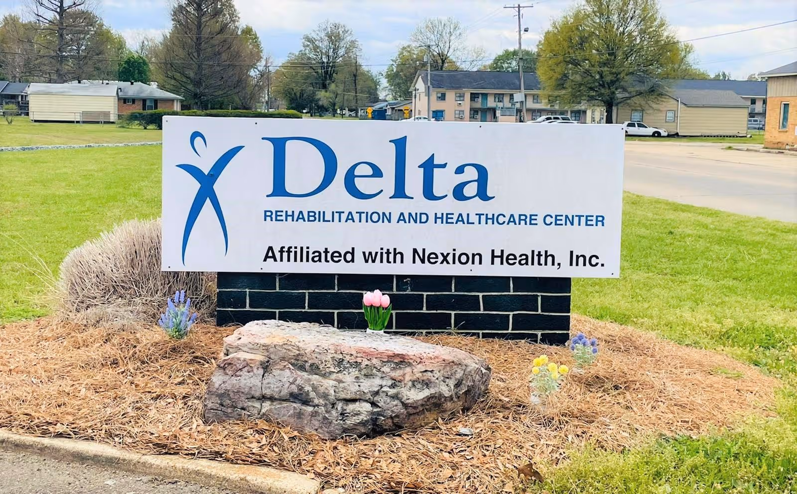 Outdoor view of a sign for Delta Rehabilitation and Healthcare Center, affiliated with Nexion Health, Inc., placed on a landscaped area with a large rock and small flowers, with residential buildings and trees in the background.