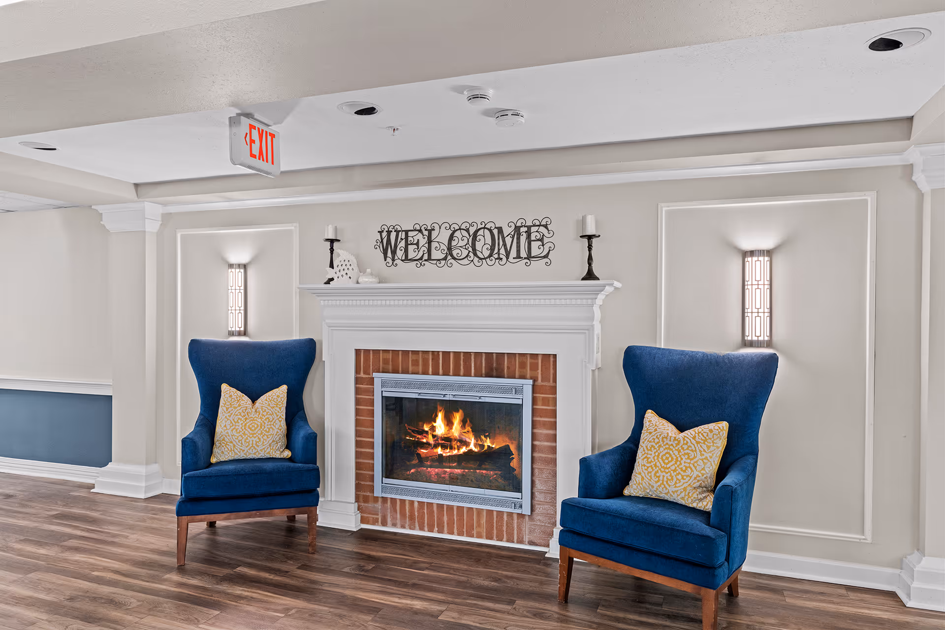 A cozy interior seating area with a lit fireplace in the center. Two blue upholstered armchairs with yellow patterned pillows are placed on either side of the fireplace. Above the fireplace is a decorative metal sign that reads 'WELCOME'. The walls are light-colored with decorative molding and wall sconces providing soft lighting. The floor is wood with a warm tone.
