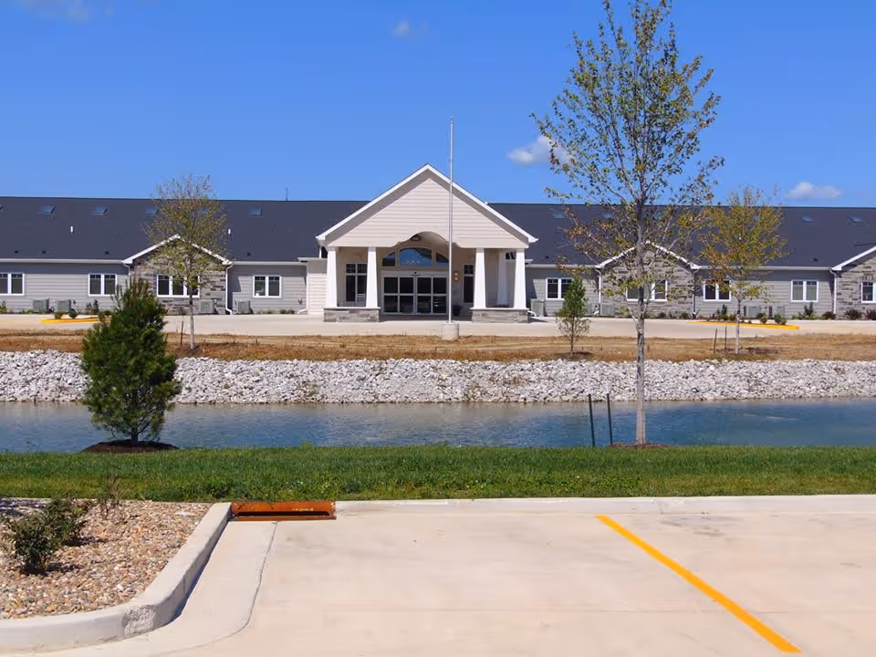Front exterior view of a single-story memory care facility building with a peaked entrance, surrounded by a small pond, trees, and a parking lot in the foreground under a clear blue sky.