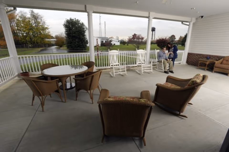 Covered outdoor patio area with wicker chairs and a round glass table, white rocking chairs along the railing, and two people sitting and standing near the corner. The background shows green grass, trees, and a water tower in the distance under a cloudy sky.