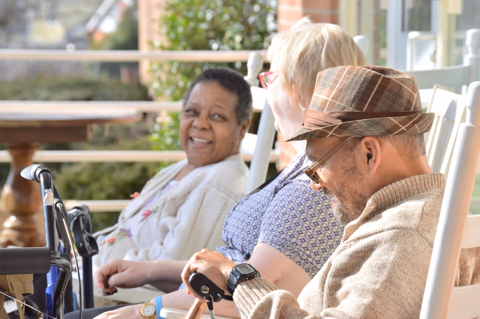 Three elderly people sitting on white rocking chairs on a porch or patio, enjoying a sunny day. One woman is smiling and looking towards the camera, another woman is looking at the man who is wearing a plaid hat and glasses, holding a cane.