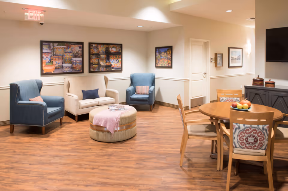 A warm, well-lit common lounge with armchairs, a loveseat, an ottoman, and a round dining table on wood flooring.