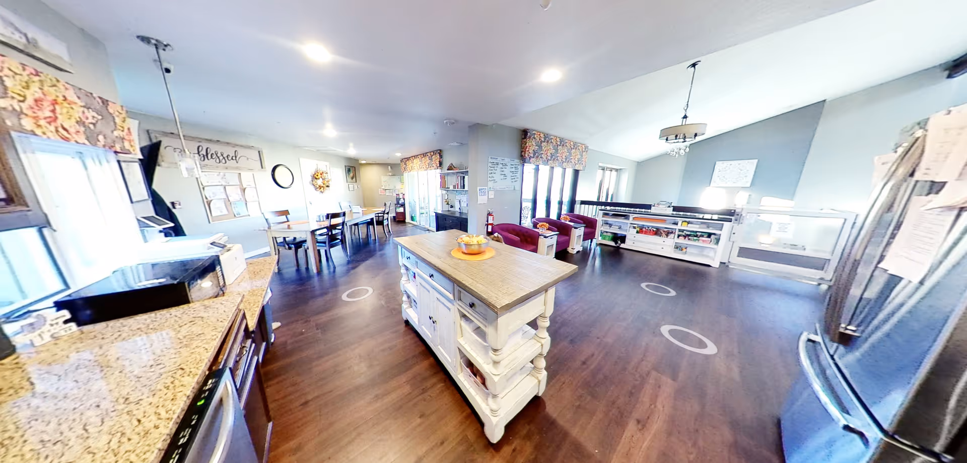 Spacious communal kitchen and dining area with a central island, tables, seating and appliances in a senior living facility.