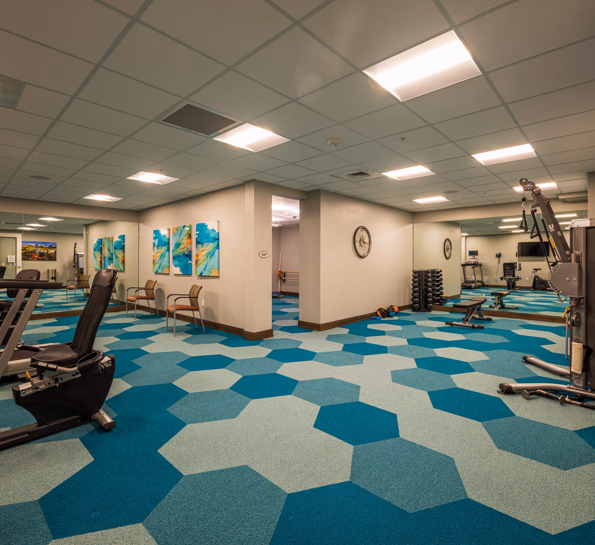 A fitness room with exercise equipment including a recumbent bike, weight rack, and multi-functional gym machine. The room has a patterned blue and teal carpet, mirrored walls, bright ceiling lights, and colorful abstract paintings on the wall. Two chairs are placed against the wall near the paintings.