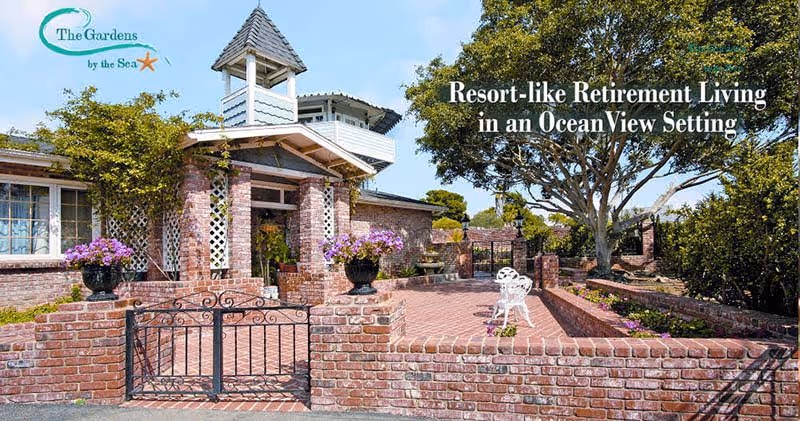 Exterior view of a brick building with a small tower and a gated entrance, surrounded by plants and flowers under a clear blue sky. There is a white bench and a large tree in the courtyard area.