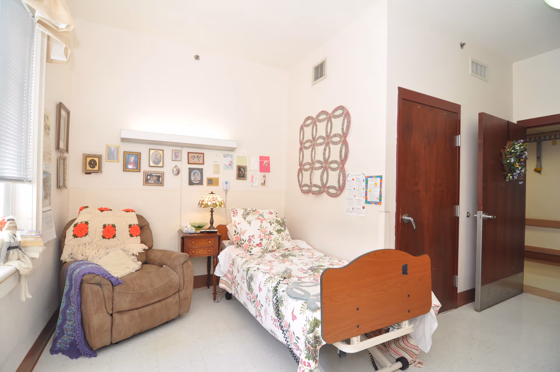 A small, cozy room in a senior living facility with a single bed covered in floral bedding, a brown recliner chair with crocheted blankets, a wooden nightstand with a decorative lamp, and several framed pictures and artwork on the walls. The room has white walls, a window with blinds, and two wooden doors, one of which is open showing a hallway.