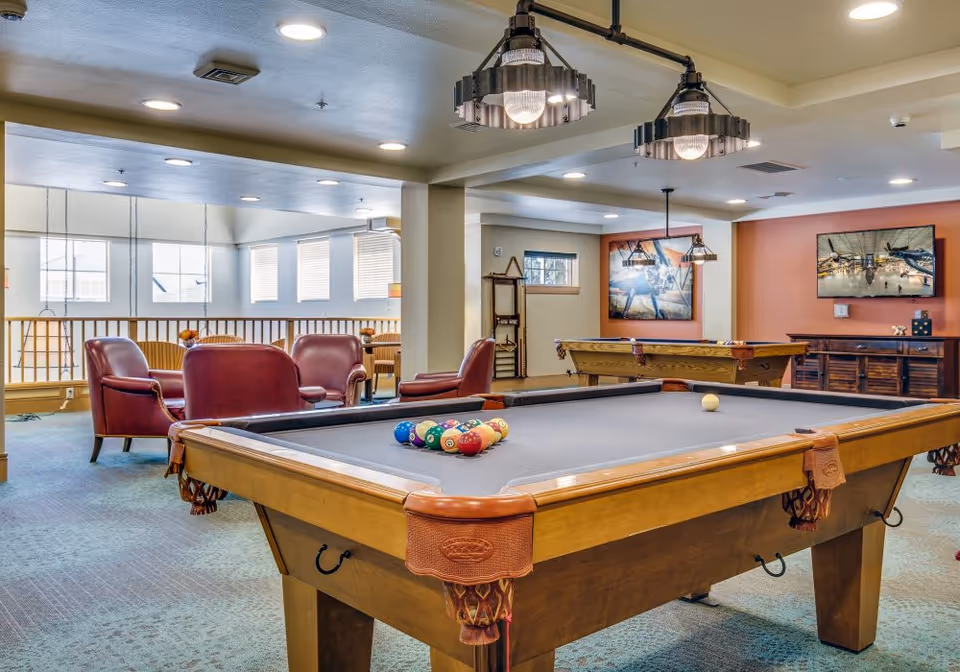 Interior view of a senior living facility game room featuring two pool tables with balls arranged on one, several red leather armchairs and sofas, hanging industrial-style light fixtures, framed artwork on the walls, and large windows letting in natural light.