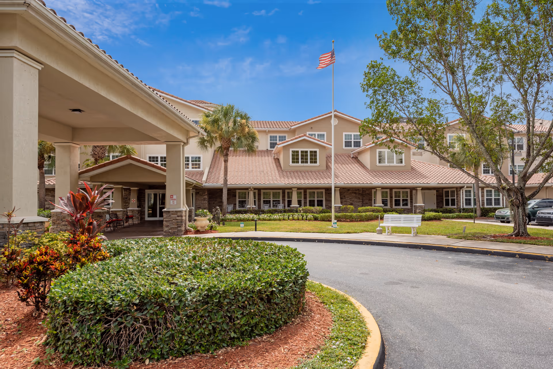 Exterior view of a senior living facility with a circular driveway, landscaped bushes, palm trees, an American flag on a flagpole, and a white bench near the entrance under a clear blue sky.