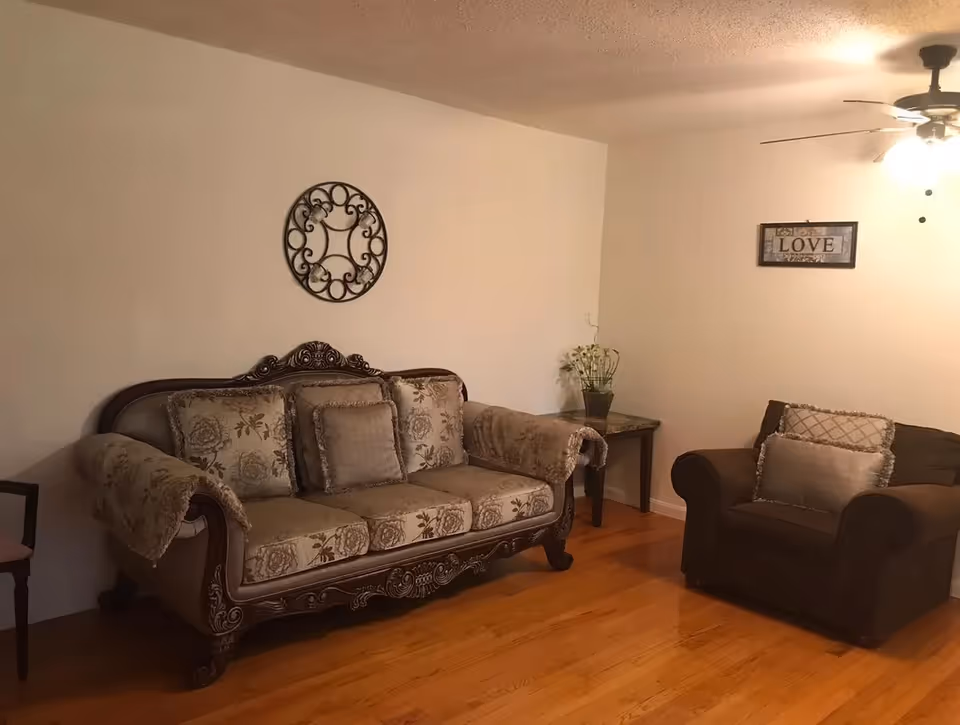 A living room with an ornate upholstered sofa and matching armchair on hardwood floors, a side table with a plant, and wall decorations.