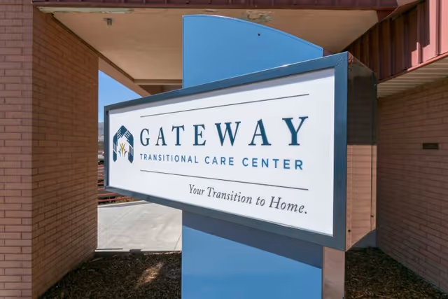 A freestanding entrance sign reading "GATEWAY Transitional Care Center — Your Transition to Home" mounted on a blue post outside the building.