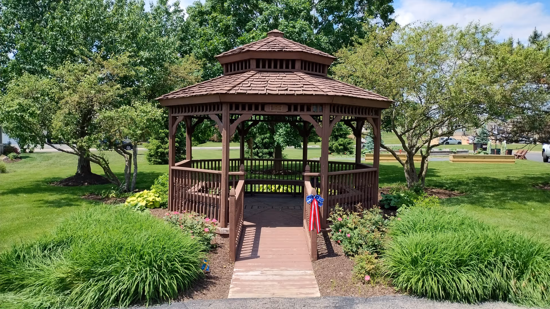 A wooden gazebo with a shingled roof is situated in a well-maintained garden area with green grass, bushes, and trees. The gazebo has a small ramp leading up to it, decorated with a red, white, and blue ribbon. The sky is partly cloudy and the surrounding area is lush and green.