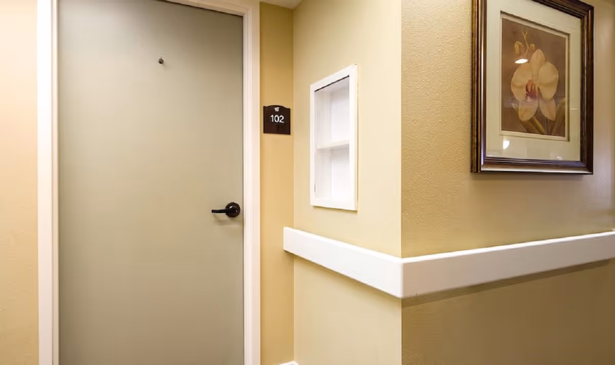 A hallway corner in a senior living facility with a closed beige door labeled 102, a white recessed wall shelf, and a framed floral painting on the wall.