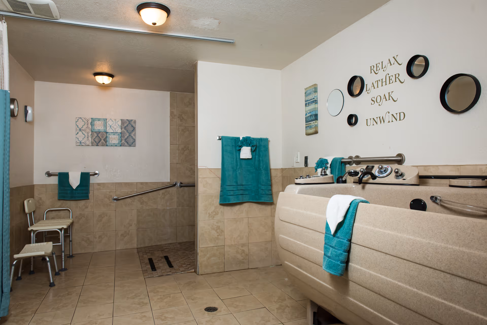 A spacious bathroom with beige tiled floors and walls. There is a large beige bathtub with control knobs and a teal towel hanging on it. On the wall above the bathtub, there are decorative mirrors and a sign that reads 'RELAX LATHER SOAK UNWIND'. The bathroom also features a walk-in shower area with grab bars, a shower chair, and teal towels hanging on the walls. The lighting consists of ceiling-mounted fixtures.